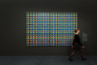 Das Bild zeigt ein abstraktes Kunstwerk mit einem Gittermuster aus bunten Rechtecken in Gelb, Blau, Rot, Grün und anderen Farben, das an einer dunklen Wand in einem Ausstellungsraum hängt. Eine Person läuft unscharf vor dem Kunstwerk vorbei. Neben dem Bild befindet sich eine Beschreibungstafel.