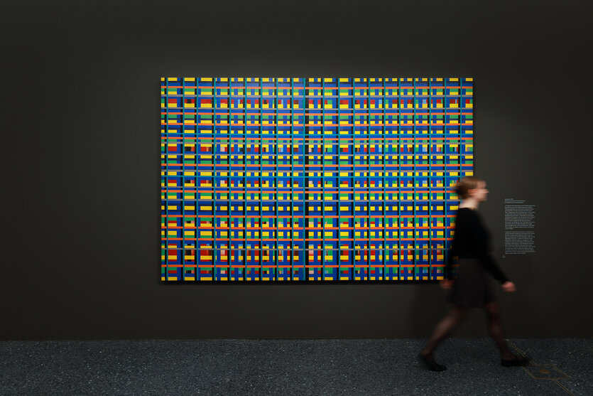 Das Bild zeigt ein abstraktes Kunstwerk mit einem Gittermuster aus bunten Rechtecken in Gelb, Blau, Rot, Grün und anderen Farben, das an einer dunklen Wand in einem Ausstellungsraum hängt. Eine Person läuft unscharf vor dem Kunstwerk vorbei. Neben dem Bild befindet sich eine Beschreibungstafel.