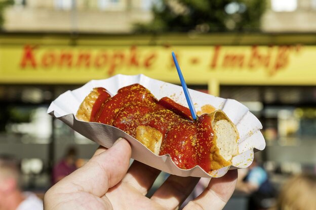EIne Schale Currywurst mit Pieker vor einem Imbiss in der Hand