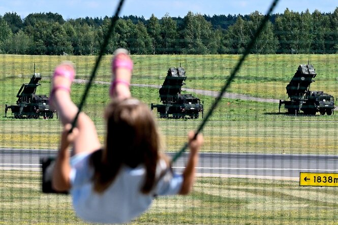 Patriot-Raketenabwehrsysteme stehen auf einer grünen Wiese, im Vordergrund schaukelt ein Mädchen
