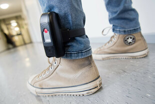 Ein Fuß mit abgetragenen Converse-Sneakern und einer schwarzen elektronischen Fußfessel am Bein in einem Flur