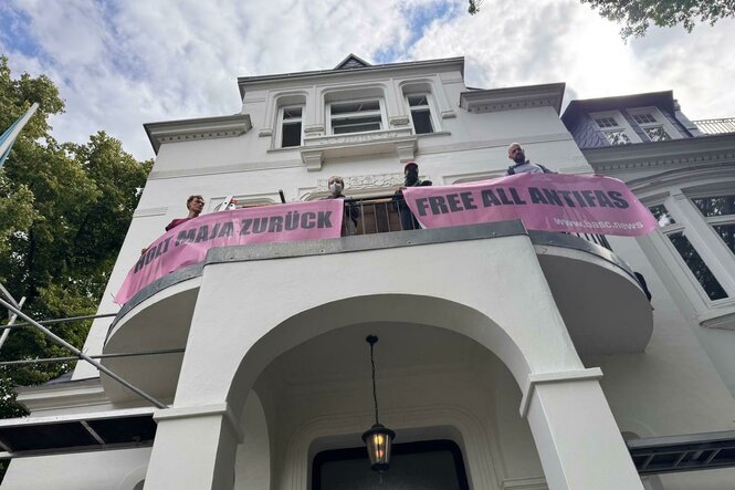 Ein Gebäude mit einem Balkon, auf dem Personen stehen und zwei rosa Banner halten. Die Banner tragen die Aufschriften "HOLT MAJA ZURÜCK" und "FREE ALL ANTIFAS" sowie eine Webadresse. Das Gebäude hat eine klassische Architektur mit einem Torbogen und einer Laterne. Im Hintergrund sind Bäume und ein blauer Himmel sichtbar.