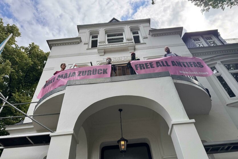 Ein Gebäude mit einem Balkon, auf dem Personen stehen und zwei rosa Banner halten. Die Banner tragen die Aufschriften "HOLT MAJA ZURÜCK" und "FREE ALL ANTIFAS" sowie eine Webadresse. Das Gebäude hat eine klassische Architektur mit einem Torbogen und einer Laterne. Im Hintergrund sind Bäume und ein blauer Himmel sichtbar.
