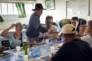 Eine Gruppe sitzt an einem Esstisch, darauf stehen Teller mit Austern und eine Flasche Wein. Eine Frau schlürft eine Auster.