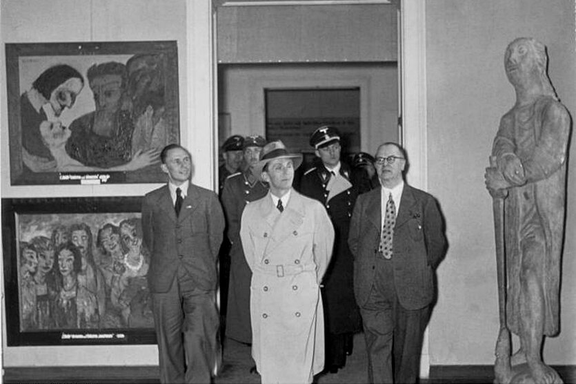 Schwarz-weiß-Foto von mehreren Männern in Anzügen und Uniformen, darunter Propagandaminister Joseph Goebbels, die vor Gemälden und einer Skulptur in einem Raum stehen