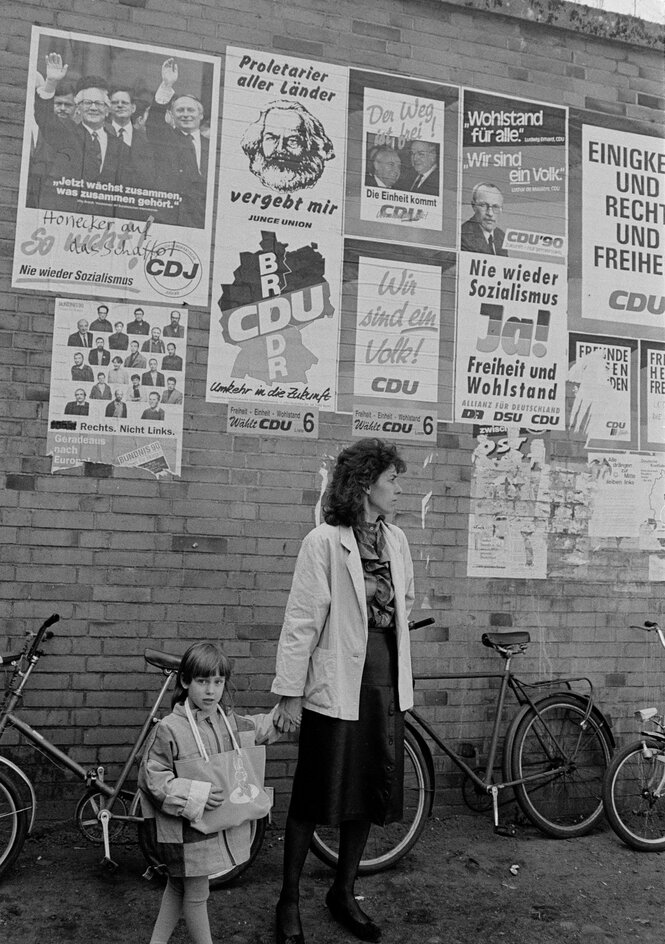 Schwarz-weiß-Foto einer Frau und eines Kinds vor einer Wand mit Plakaten