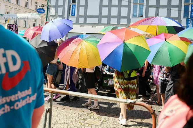 Teilnehmerinnen und Teilnehmer des CSD (Christopher Street Day) halten Schirme in Regenbogenfarben, während sie unter Politzeischutz am Marktplatz vorbeigehen, auf dem die AfD ein Sommerfest abhält