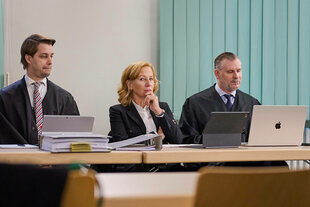 Eine Frau sitzt zwischen zwei Männern an einem Tisch im Gerichtssaal