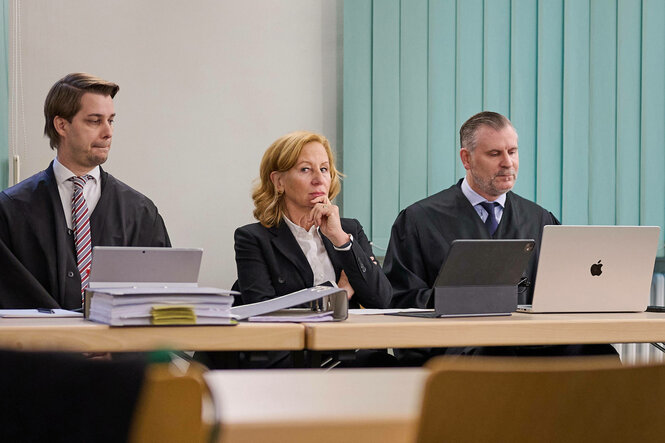 Eine Frau sitzt zwischen zwei Männern an einem Tisch im Gerichtssaal
