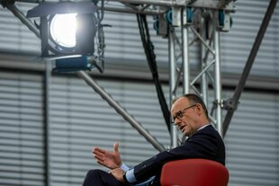 Kanzler Merz sitzt in einem roten Sessel unter einem Scheinwerfer und gestikuliert