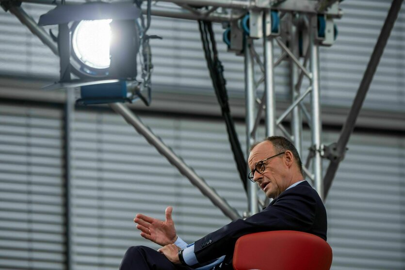Kanzler Merz sitzt in einem roten Sessel unter einem Scheinwerfer und gestikuliert