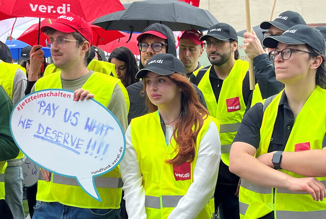Protestierende mit gelben Warnwesten halten ein Schild hoch, auf dem steht: "Pay us what we deserve"