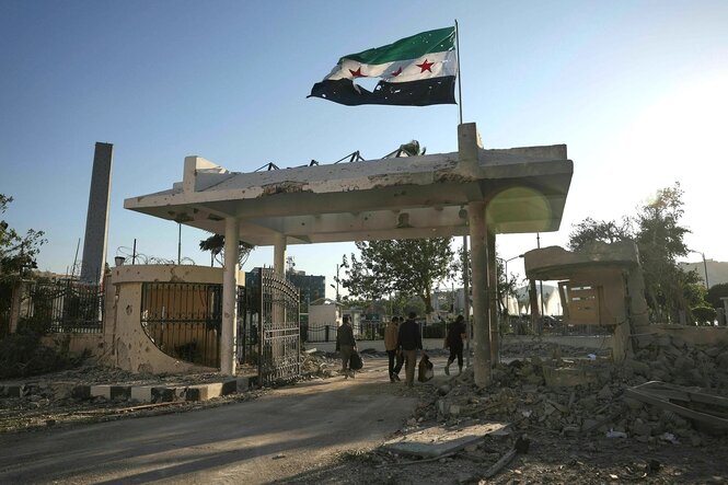 Sicherheitskräfte gehen mit Taschen in der Hand durch ds Tor des Verteidigungsministeriums auf dem eine zerfetzte syrische Flagge weht. Spuren der Zerstörung, Schutt liegt herum