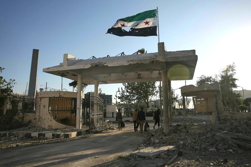Sicherheitskräfte gehen mit Taschen in der Hand durch ds Tor des Verteidigungsministeriums auf dem eine zerfetzte syrische Flagge weht. Spuren der Zerstörung, Schutt liegt herum