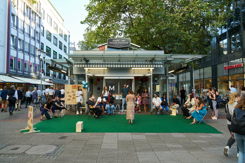 Das Umzu in der Bremer Innenstadt bei der Eröffnung eines Projekts für Mehrsprachigkeit, Moyo. Auf einem grünen Teppich rund um den Glaskasten am Hanseatenhof sitzen und stehen diverse Leute, in der Mitte eine Frau im Kleid. Als sogenannter Dritter Ort sollte das Umzu Begegnungen ohne Konsum ermöglichen