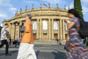 Ein großes, klassizistisches Gebäude aus Sandstein oder hellem Stein, das Opernhaus Stittgart, dominiert das Bild. Es verfügt über eine Säulenfront, mehrere Fenster und Statuen entlang der Dachlinie, auf der eine deutsche Flagge weht. Im Vordergrund sind mehrere Personen unscharf und in Bewegung festgehalten, was auf eine belebte öffentliche Szene hindeutet. Der Himmel ist leicht bewölkt und das Licht wirkt wie am späten Nachmittag.