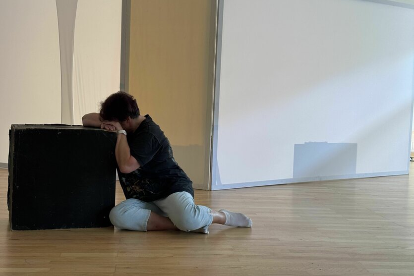 Eine Person sitzt auf hellem Laminatboden, Oberkörper und Kopf an eine schwarze Kiste angelehnt