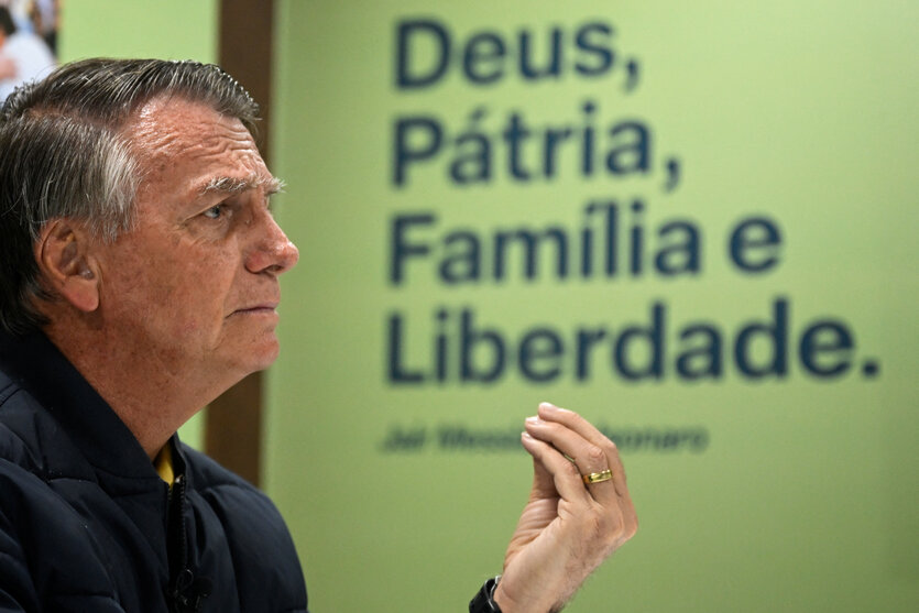 Jair Bolsonaro, im Seitenprofil, macht vor einem grünen Plakat mit der Aufschrift Deus, Patria, Familia e Liberdade eine Geste mit seiher Hand