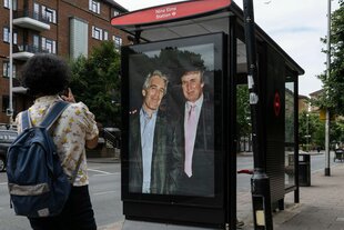 Die kurze Seite einer Bushaltestelle. Dort hängt an dem Ort, an dem sonst Werbeplakate hängen, ein riesiges Bild von Epstein und Trump, die in Anzug eng nebeneinanderstehen. Ein Mensch steht vor dem Plakat und schaut es an und macht ein Foto mit seinem Handy.