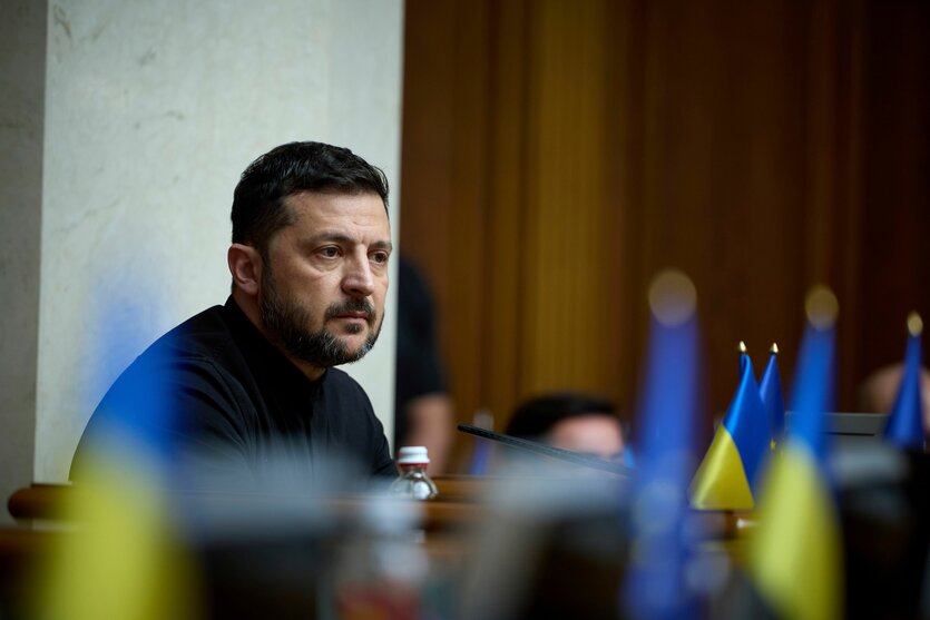 Selenskyj sitzt an einem Pult, vor ihm eine Reihe ukrainischer Flaggen