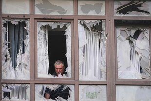 Eine Fensterfront, alle Fenster sind kaputt, durch eines der Fenster schaut grimmig eine Person