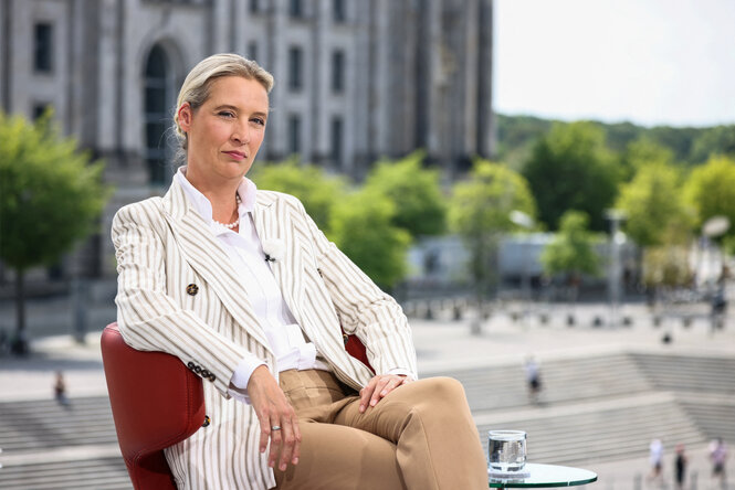 Alice Weidel sitzt auf einem Stuhl, im Hintergrund das Berliner Reichstagsviertel