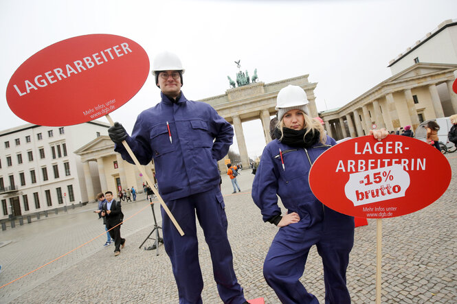Ein Mann und eine Frau in blauer Arbeitskleidung stehen vor dem Brandburger Tor. Die Frau hält ein Schild mit der Aufschrift "minus 15 Prozent brutto".