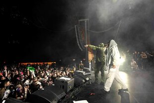 Zwei Mitglieder der BandKneecap stehen auf der Bühne, einer trägt ein Palituch auf dem Kopf und singt, im Publikum wird ein palästinensische Flagge hochgehalten