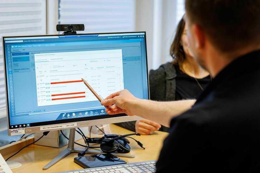 Ein Arzt sitzt am Rechner und zeigt mit dem Stift auf die Daten einer Patienten, die vor ihm sitzt, aber fast vollständig von ihm verdeckt wird