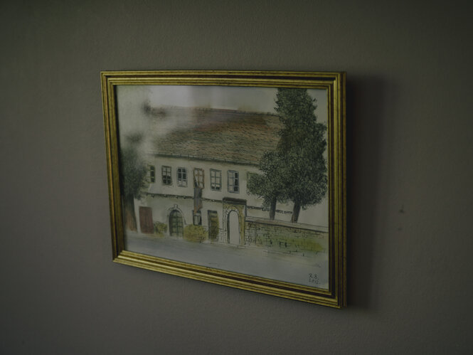 Eine Zeichnung eines Hauses hängt in einem goldenen Bilderrahmen an der grauen Wand