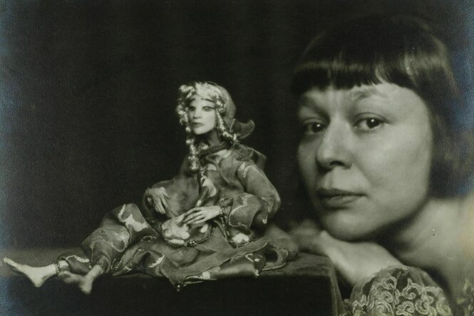Eine zierliche Puppe sitzt auf einem Podest, neben ihr stützt die Künstlerin Lotte Pritzel ihr Kinn auf die Hand und schaut den Betrachtenden an