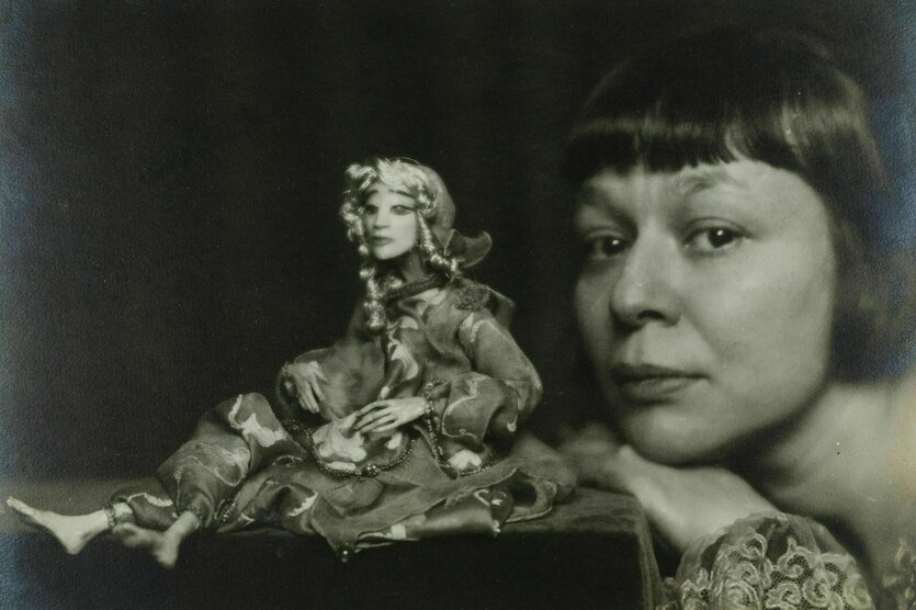 Eine zierliche Puppe sitzt auf einem Podest, neben ihr stützt die Künstlerin Lotte Pritzel ihr Kinn auf die Hand und schaut den Betrachtenden an
