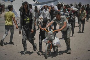 Zwei Männer haben einen Sack Mehl auf eine Kinderfahrrad geladen, das sie gemeinsam mitdem Kind schieben. Hinter ihnen sind Menschen zu erkennen, von denen manche auch einen Sack Mehl transportieren