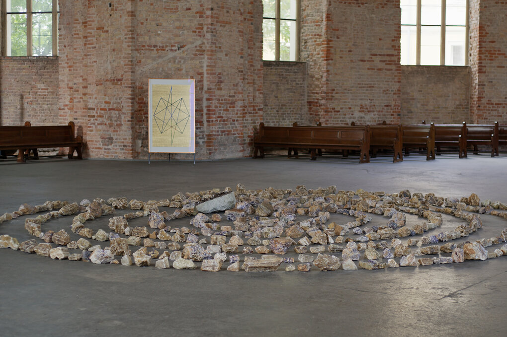 Bilder von Engeln und Sternenkonstellationen lehnen an der kargen Backsteinwand einer Kiche, dazu ein Labyrinth aus Steinen auf dem Boden