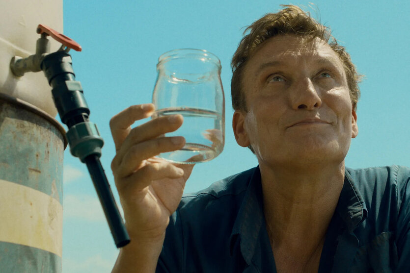 Filmstill zeigt den Schauspieler Oliver Masucci als Uri, er hält ein Glas Wasser in der Hand, neben ihm ein Wasserfass mit Hahn, er schaut nachdenklich nach oben