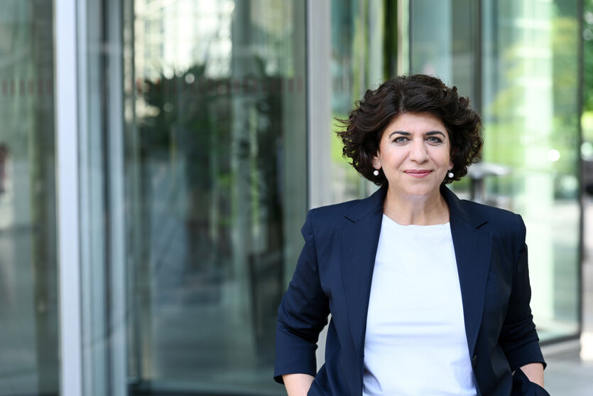 Berivan Aymaz steht vor einer Glaswand, sie trägt ein dunkles Jacket und ein weisses T-Shirt, sie lächelt freundlich