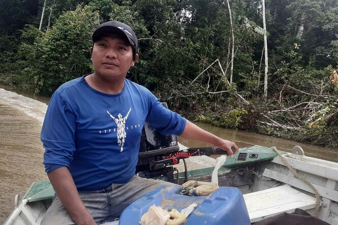 Indigener mit Käppi und blauem T-Shirt im Motorboot, einen Fluss entlang fahrend