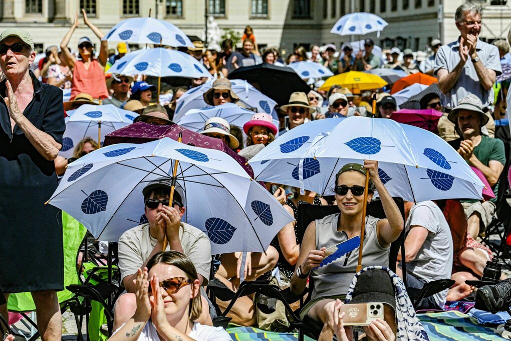 Besucher der Veranstaltung "Staatsoper für alle" sitzen auf dem Bebelplatz mit Sonnenschirmen, auf Decken, mitgebracuhten Stühlen, manache applaudieren stehend