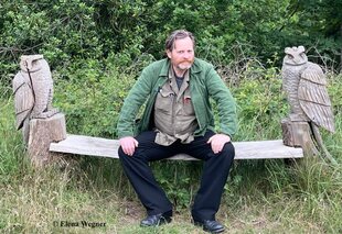 Bernhard Marsch sitzt auf einer Wiese auf der Holzbank gerahmt von zwei Eulen
