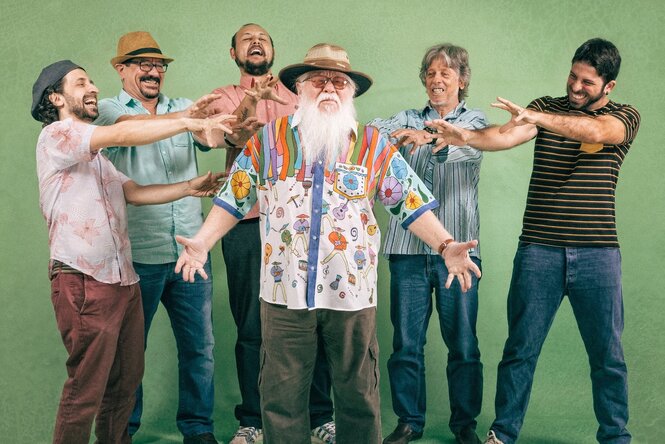 Der Musiker Hermeto Pascoal trägt einen langen weißen Bart, einen Hut und ein weißes Hemd. Er ist von fünf Bandmitgliedern umgeben, die zelebrierend ihre Arme auf ihn richten.