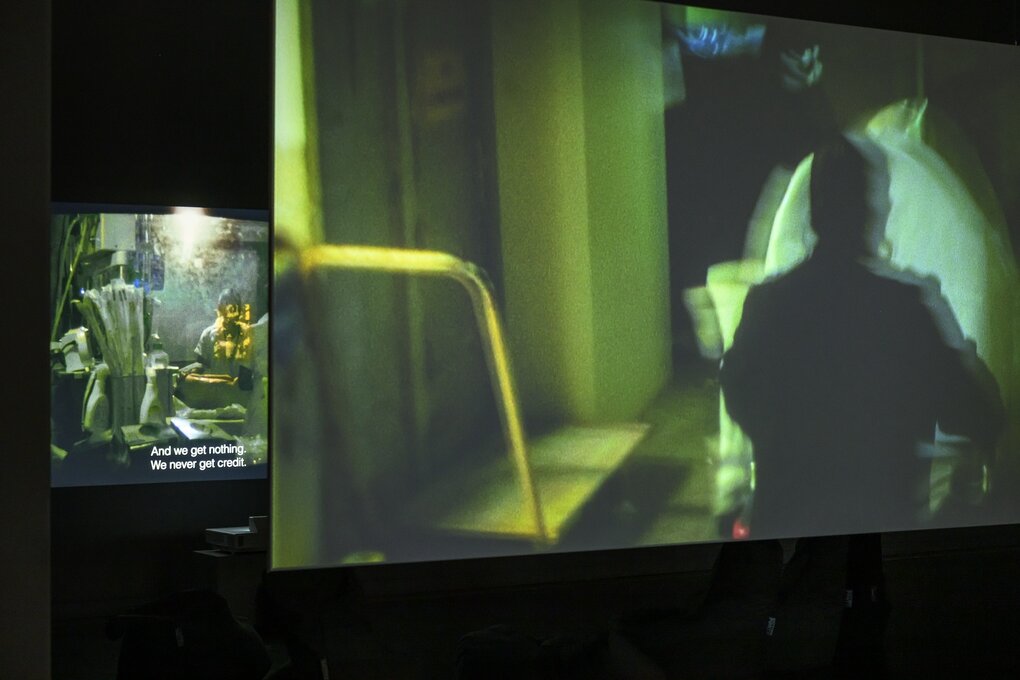 Blick in die Betonhalle des silent green Kulturquartier. Auf zwei Leinwänden sind Ausschnitte aus einem Film zu sehen, in grünen Farben gehalten, zeigen die Bilder einen Operationssaals (Leinwand hinten links) und die Umrisse eines Arztes (Leinwand vorne rechts im Bild).