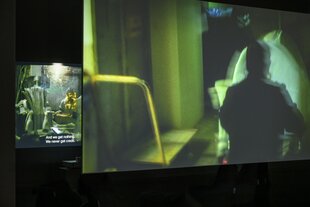 Blick in die Betonhalle des silent green Kulturquartier. Auf zwei Leinwänden sind Ausschnitte aus einem Film zu sehen, in grünen Farben gehalten, zeigen die Bilder einen Operationssaals (Leinwand hinten links) und die Umrisse eines Arztes (Leinwand vorne rechts im Bild).