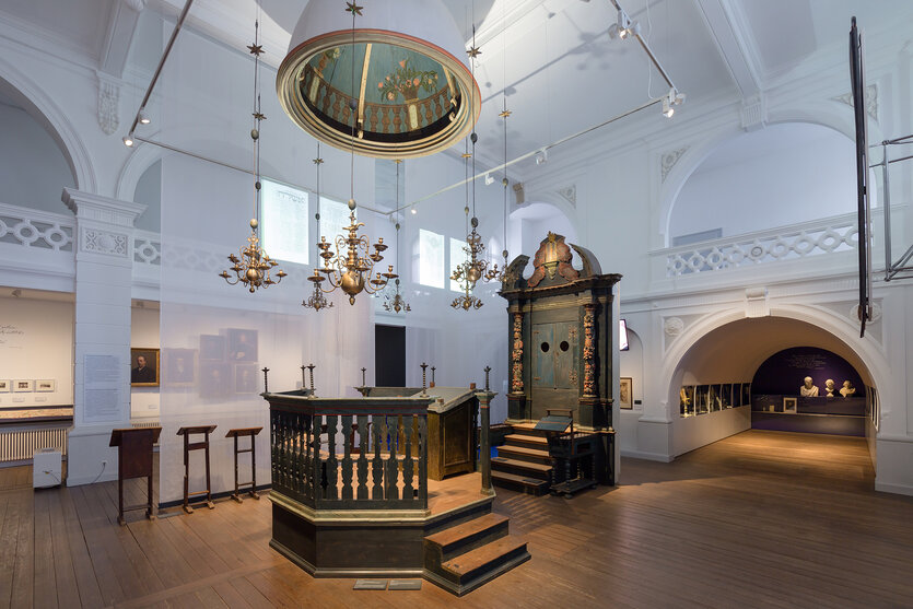 Historischer Raum mit hoher, weißer Decke, Bögen und Ornamenten. Im Zentrum ein großer, verzieter Holzaltar mit Stufen, umgeben von einem geschnitzten Gehäuse. Darüber ein runder Kronleuchter mit floralen Motiven, ergänzt durch kleinere Kerzenleuchter. Wände mit Porträts und Büsten, dunkler Holzfußboden, modernes Licht.