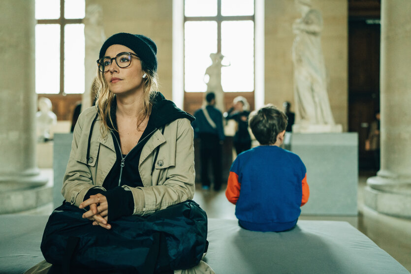 Eine Frau mit einer schwarzen Tasche auf ihrem Schoß in einem Museum. Ein Junge in einem blauen Pulli sitzt mit dem Rücken zu ihr