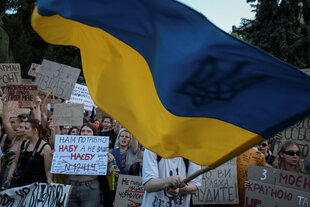 Demonstrierende halten Protestschilder und schwenken eine große ukrainische Flagge