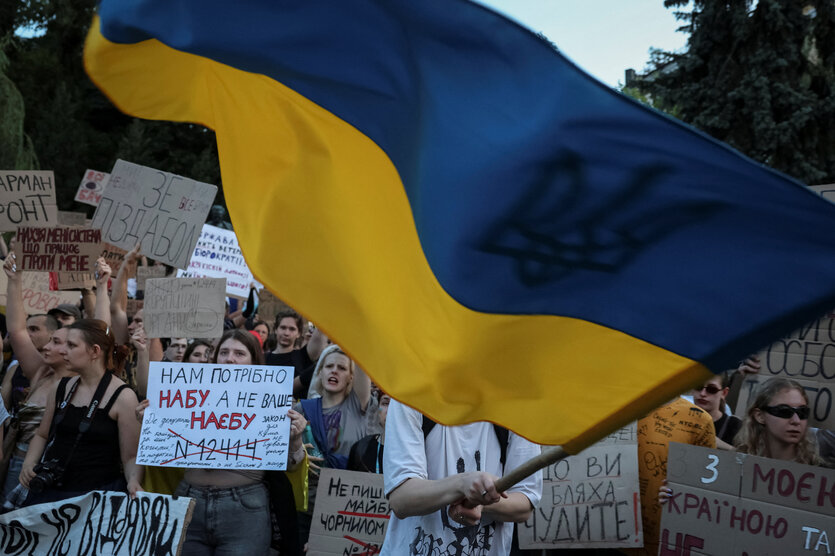 Demonstrierende halten Protestschilder und schwenken eine große ukrainische Flagge