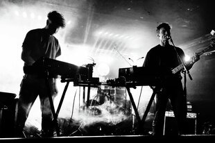 Schwarz-Weiß-Aufnahme des Duos Isolationsgemeinschaft bei einem Live-Auftritt. Die Musiker stehen mit Keyboard und Gitarre auf der Bühne, die in Nebel getaucht ist.