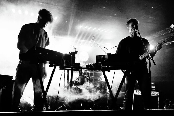 Schwarz-Weiß-Aufnahme des Duos Isolationsgemeinschaft bei einem Live-Auftritt. Die Musiker stehen mit Keyboard und Gitarre auf der Bühne, die in Nebel getaucht ist.