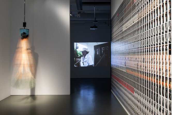 In einem Ausstellungsraum ist ein mit einem Motor betriebener, im Kreis aufgehangener Vorhang aus Bambusketten zu sehen, auf den ein Gesicht aufgedruckt ist. Das Kunstwerk hängt von der Decke und befindet sich im Drehmoment. Die Wand daneben überzieht ein Fotocollage. Im Hintergrund zeigt eine Videoarbeit einen Postboten bei der Arbeit.