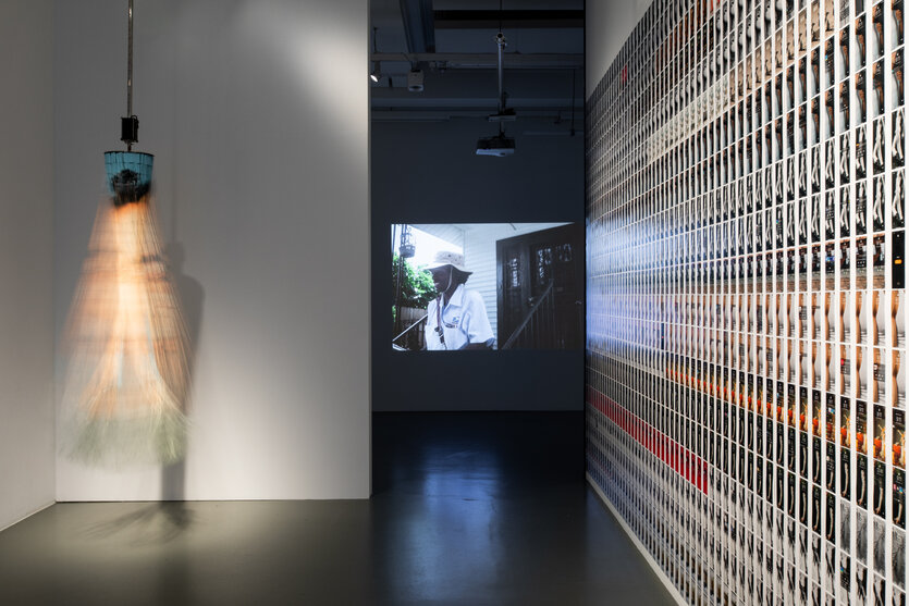 In einem Ausstellungsraum ist ein mit einem Motor betriebener, im Kreis aufgehangener Vorhang aus Bambusketten zu sehen, auf den ein Gesicht aufgedruckt ist. Das Kunstwerk hängt von der Decke und befindet sich im Drehmoment. Die Wand daneben überzieht ein Fotocollage. Im Hintergrund zeigt eine Videoarbeit einen Postboten bei der Arbeit.